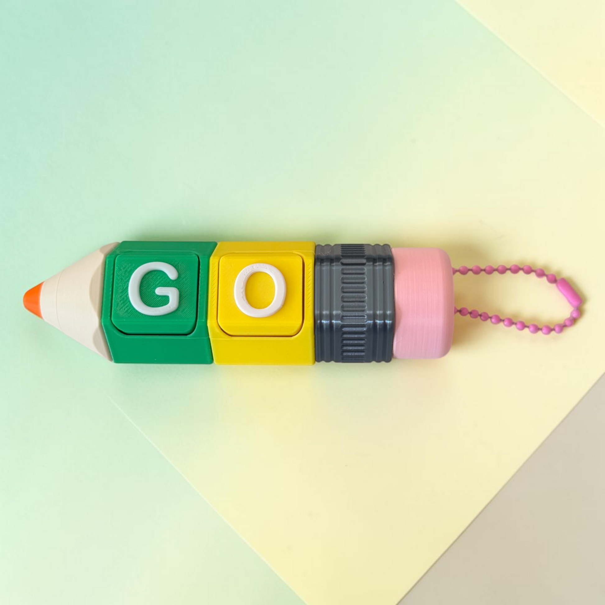 A pencil-shaped novelty fidget keychain  with colorful blocks spelling 'GO' in green and yellow, a dark gray ribbed middle section, and a pink eraser end with a pink bead chain, photographed on a pastel multicolor background.