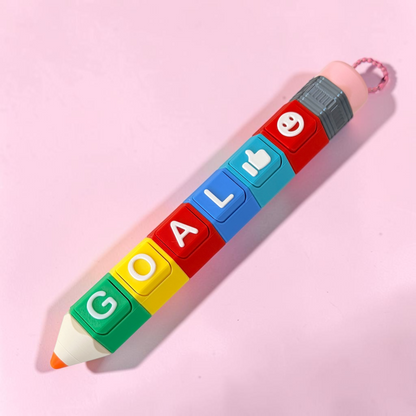 A novelty pencil shaped fidget toy with colorful lettered segments spelling 'GOAL' and emoji icons, photographed on a soft pink background.