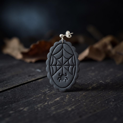 A black oval shaped like a spiderweb, hanging from a silver stud earring on a dark wooden surface with blurred dried leaves in the background.