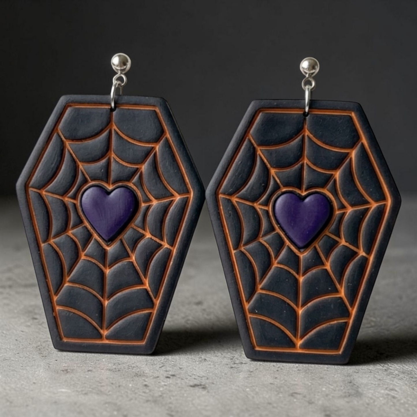 A pair of coffin-shaped earrings with a black and dark gray spiderweb pattern outlined in orange, featuring a purple heart in the center and silver-tone hooks.