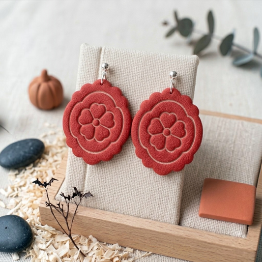 A pair of red flower-shaped drop earrings displayed on a beige fabric jewelry stand, styled with small decorative stones, a wooden tray, and eucalyptus leaves on a light neutral background.