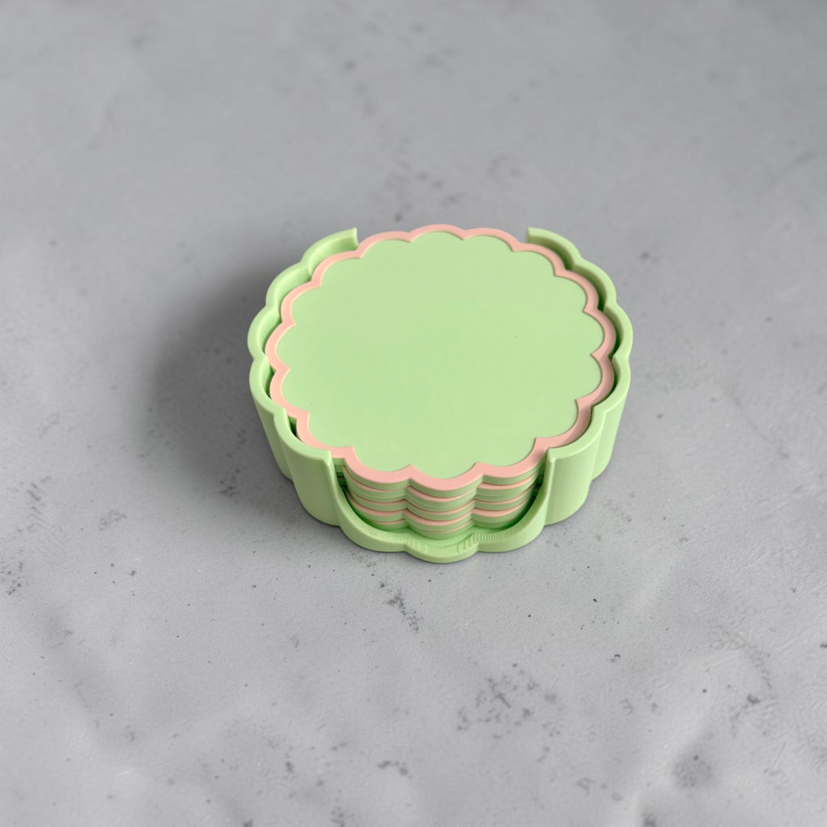 A stack of scalloped-edged coasters   in pastel green and pink, resting on a light gray marble surface.