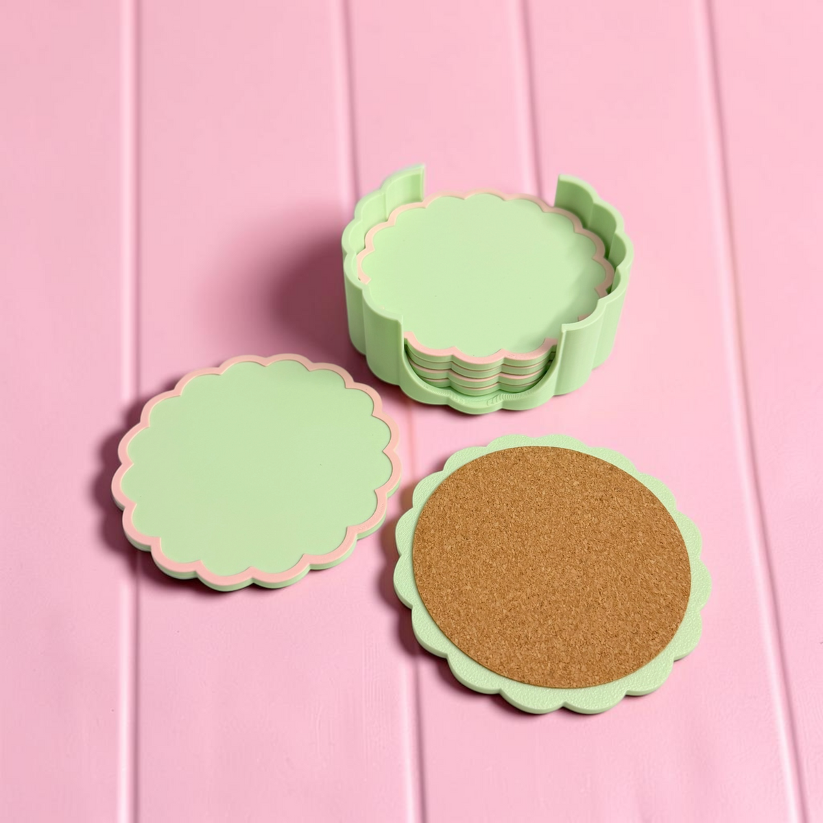 A set of round scalloped coasters in mint green with a pink inner edge, shown stacked and individually on a pink wood-plank background. One coaster is shown with a cork insert.